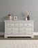 Vermont 3+2 Drawer Chest in Ivory, a modern furniture piece with multiple drawers and a clean design, placed against a minimalist wall with decorative vases and a lamp on top.