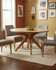Stanley Round Table (130cm), a wooden dining table with a smooth round surface and sturdy angular legs, surrounded by four gray upholstered chairs, in a cozy dining room with framed family photos on the wall.