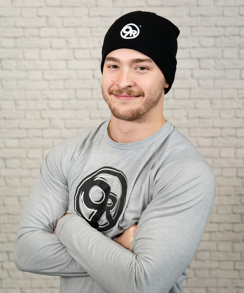 A young adult male wearing a black beanie with a white 9Round circle logo on the front center and a grey long-sleeve t-shirt with a large black 9Round circle logo on the center chest