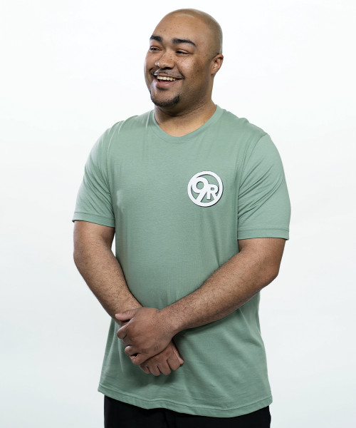 A young adult male smiling wearing a sage green t-shirt with a big white circle logo for 9Round Fitness on the upper right-hand area of the chest.