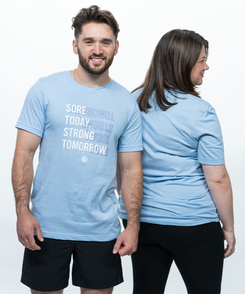 A young adult male smiling at the camera wearing a light blue unisex t-shirt that reads, "Sore Today, Strong Tomorrow," in a white font with a dark blue outline of the same text behind it. Along with a young adult female wearing a light blue shirt facing away from the camera.