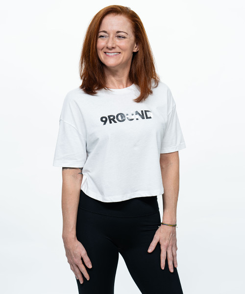 A young adult woman smiling with long red hair wearing a boxy white t-shirt that is cropped at the waist with a silver foil 9Round logo printed on the front. She is also wearing black leggings.