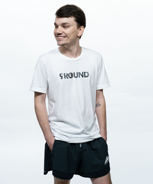 A young adult male smiling off to the side with a white unisex t-shirt that has a silver foil printed 9Round logo on the front. His hands are in the pockets of a pair of black shorts.