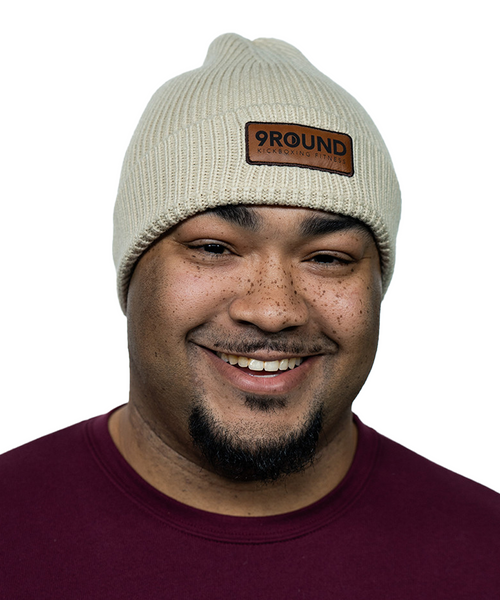 A young male wearing a natural colored knit beanie with a brown leather patch that reads 9Round