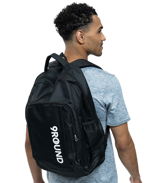 A young adult male wearing a blue t-shirt and black pants with a black backpack that has a vertically placed white 9Round logo on the front larger pocket