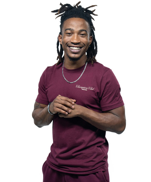 Man with dreadlocks wearing a maroon t-shirt and matching pants stands against a white background, smiling with hands clasped.