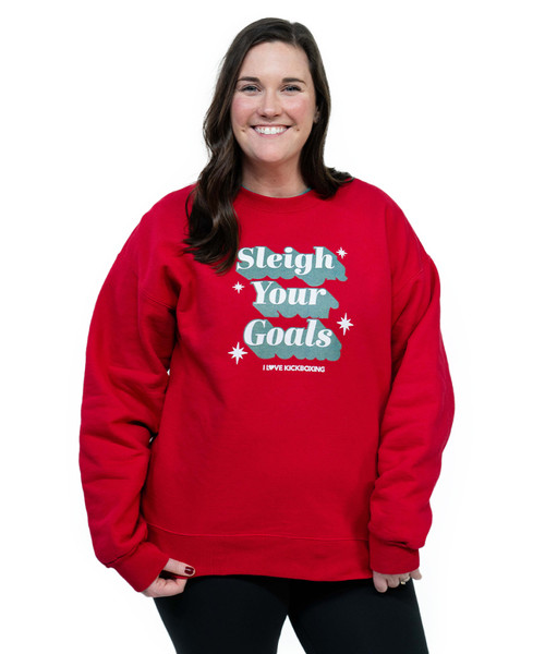 Woman wearing a red sweatshirt with the text Sleigh Your Goals and small stars, standing against a white background and smiling.