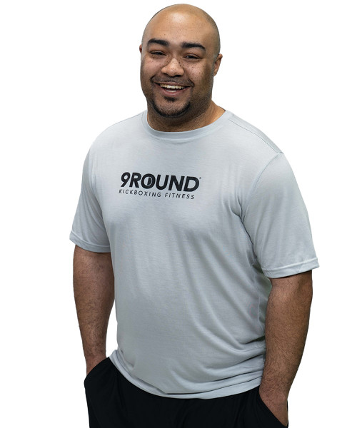 Smiling man in a light gray 9Round Kickboxing Fitness t-shirt with hands in pockets, standing against a plain white background.