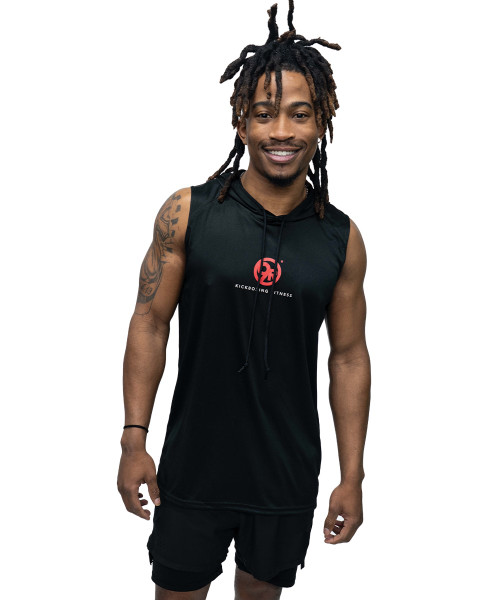 A man with dreadlocks wearing a sleeveless black hoodie and black shorts stands smiling against a white background.