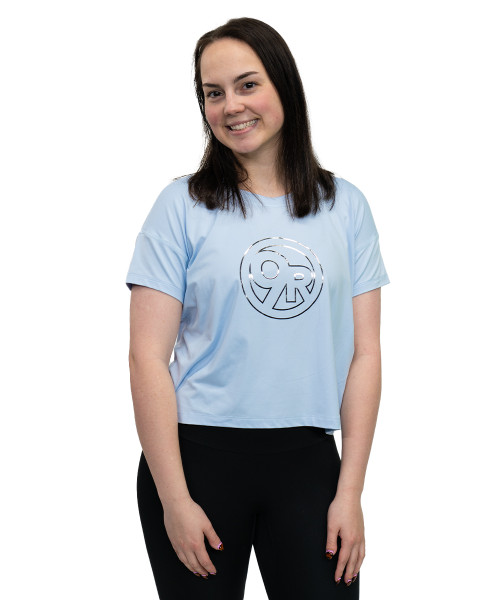 A woman with straight dark hair wearing a light blue t-shirt with a circular logo and black pants stands against a white background, smiling at the camera.