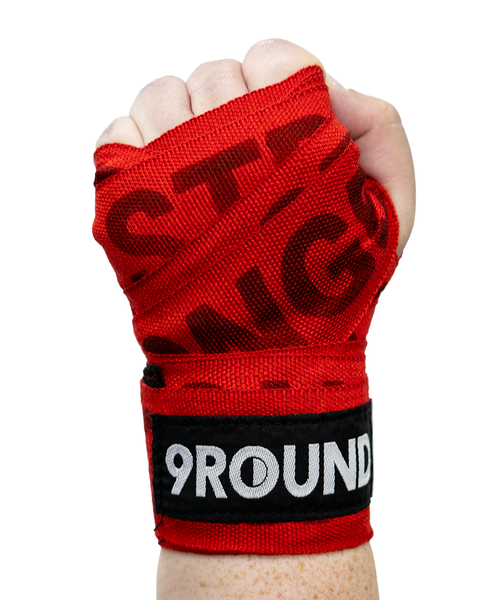 A fist wrapped in red hand wraps featuring the 9ROUND logo on a black band, isolated on a white background.