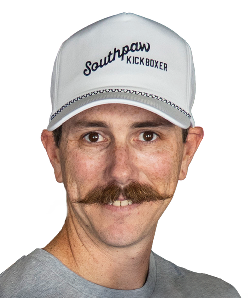 A person with a mustache wearing a white Southpaw Kickboxer cap and a gray shirt, facing the camera against a white background.