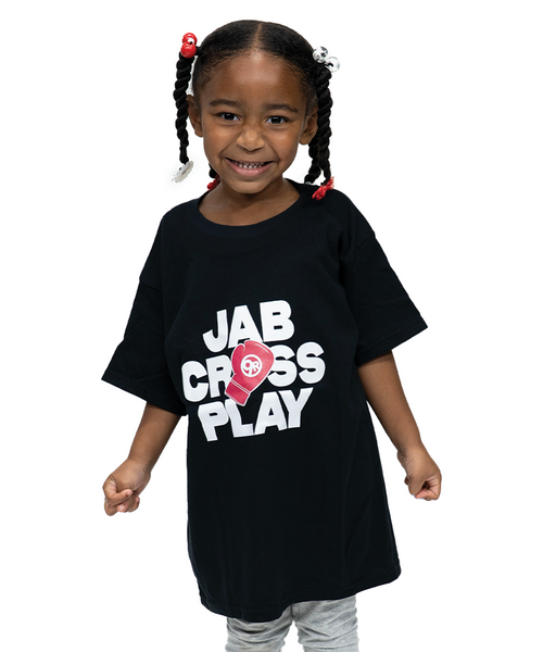 Young girl with braided hair smiles while wearing a black T-shirt that says JAB CROSS PLAY with boxing glove graphics on a white background.