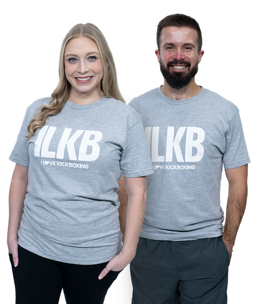 Two people wearing matching gray ILKB I Love Kickboxing t-shirts stand close together, smiling at the camera against a white background.