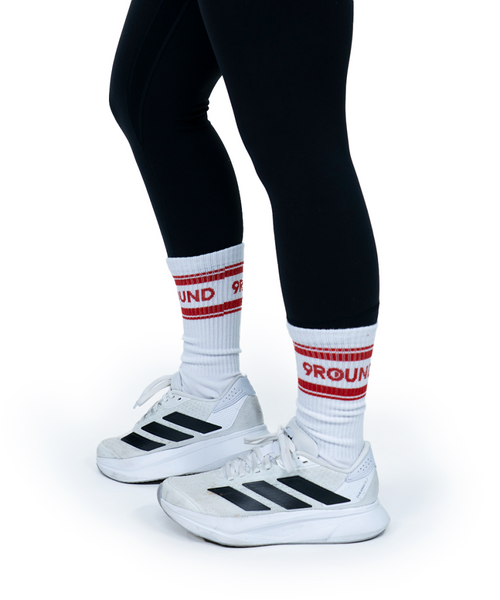 Person wearing black leggings, white athletic shoes with black stripes, and white socks with red stripes and 9ROUND text. The image is taken against a plain white background.