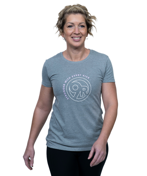 A woman with short, wavy blonde hair smiles while wearing a gray t-shirt with a circular logo and text that reads Stronger With Every Kick. She stands against a white background.