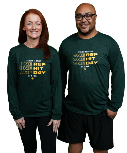 Two people stand side by side wearing matching long-sleeve shirts that read Strength is built one rep, one hit, one day at a time. Both are smiling at the camera.