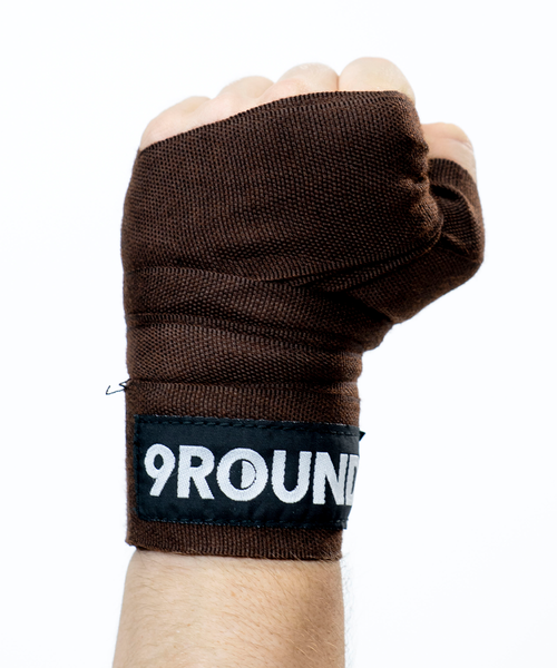 A person making a fist in the air with their hand wearing a brown boxing hand wrap that reads 9Round across the wrist 