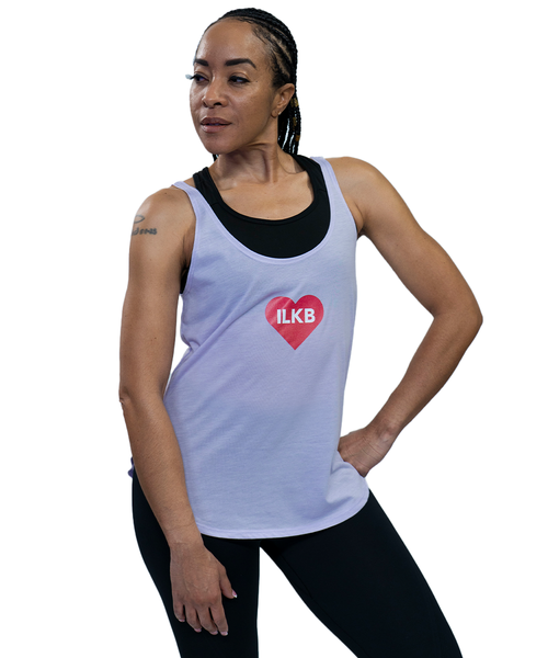 Woman in athletic wear poses with one hand on hip, wearing a white tank top with a red heart and ILKB logo, over a black sports bra and black leggings.