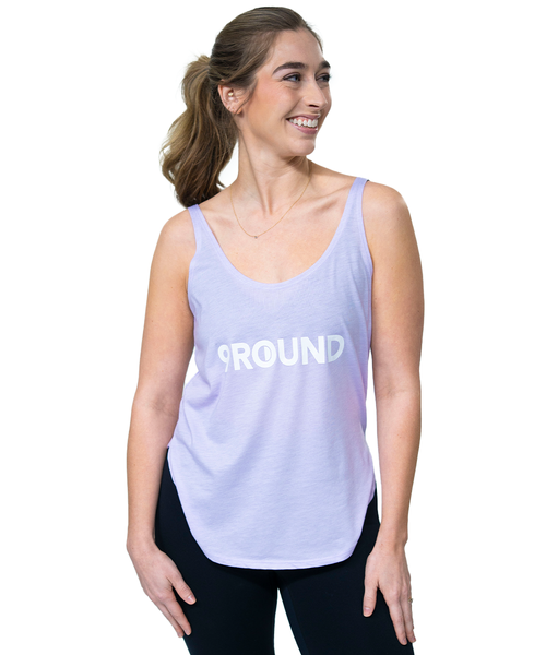 Woman wearing a light purple tank top with 9ROUND printed on the front, standing against a white background and smiling while looking to the side.
