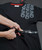 An image of someone cutting black t-shirt with three grey 9Round logos printed in a modern design on the upper right-hand shoulder. They are cutting along a provided crop line with tape marking where to cut.