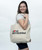 A woman with brown hair with a canvas tote bag on her shoulder looking off to the side. The tote bag has a navy and red I Love Kickboxing logo.
