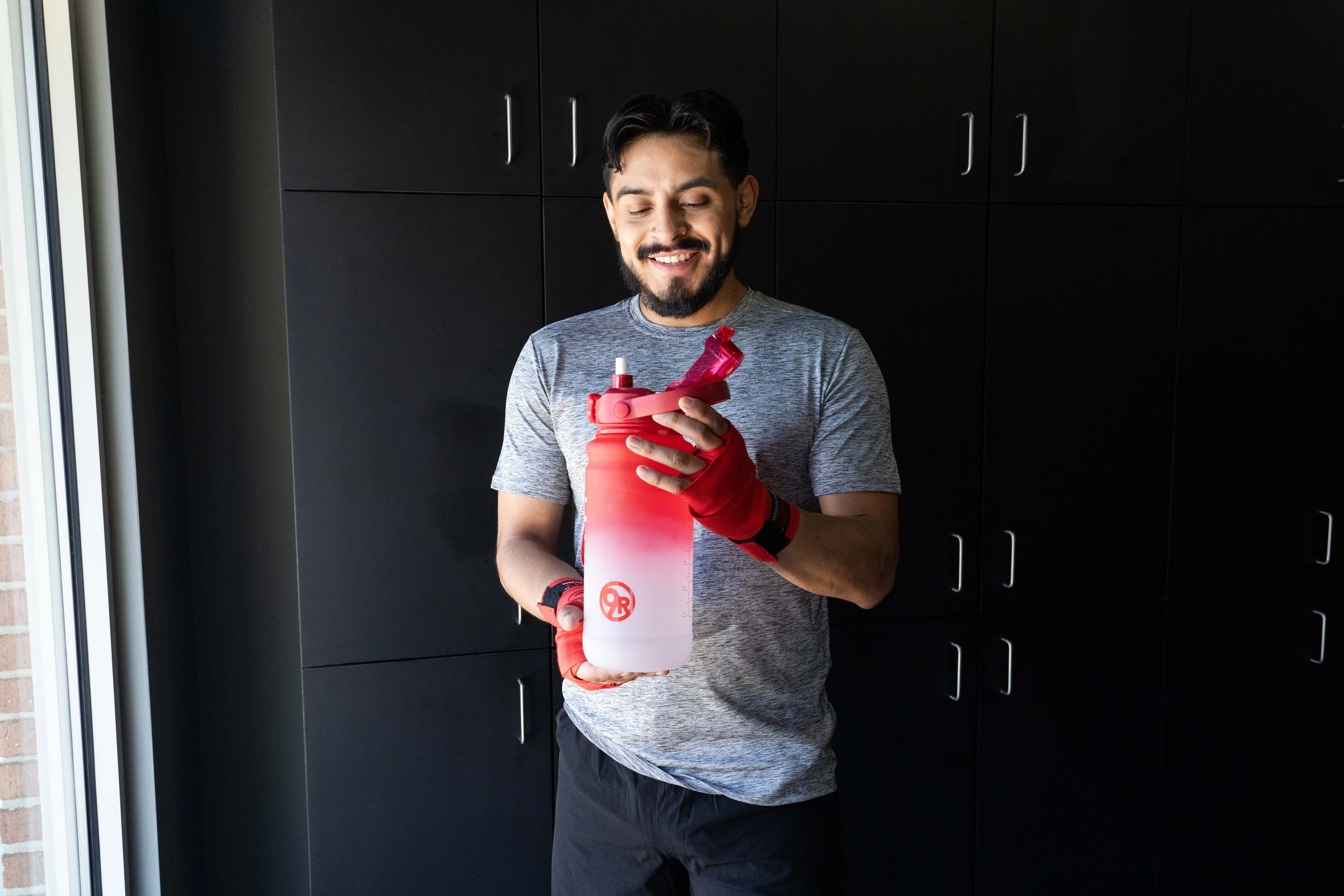 A man with dark black hair in a grey t-shirt and black shorts holding a red and white water bottle with a red 9Round circle logo standing in front of a set of black lockers.