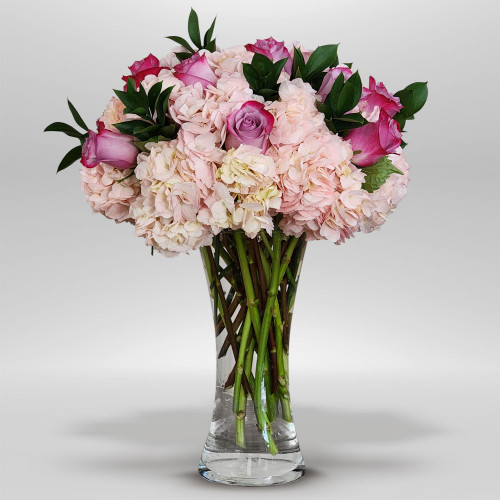 is a soft and elegant floral arrangement featuring blush hydrangeas, pink roses, and fresh greenery, arranged in a clear glass vase