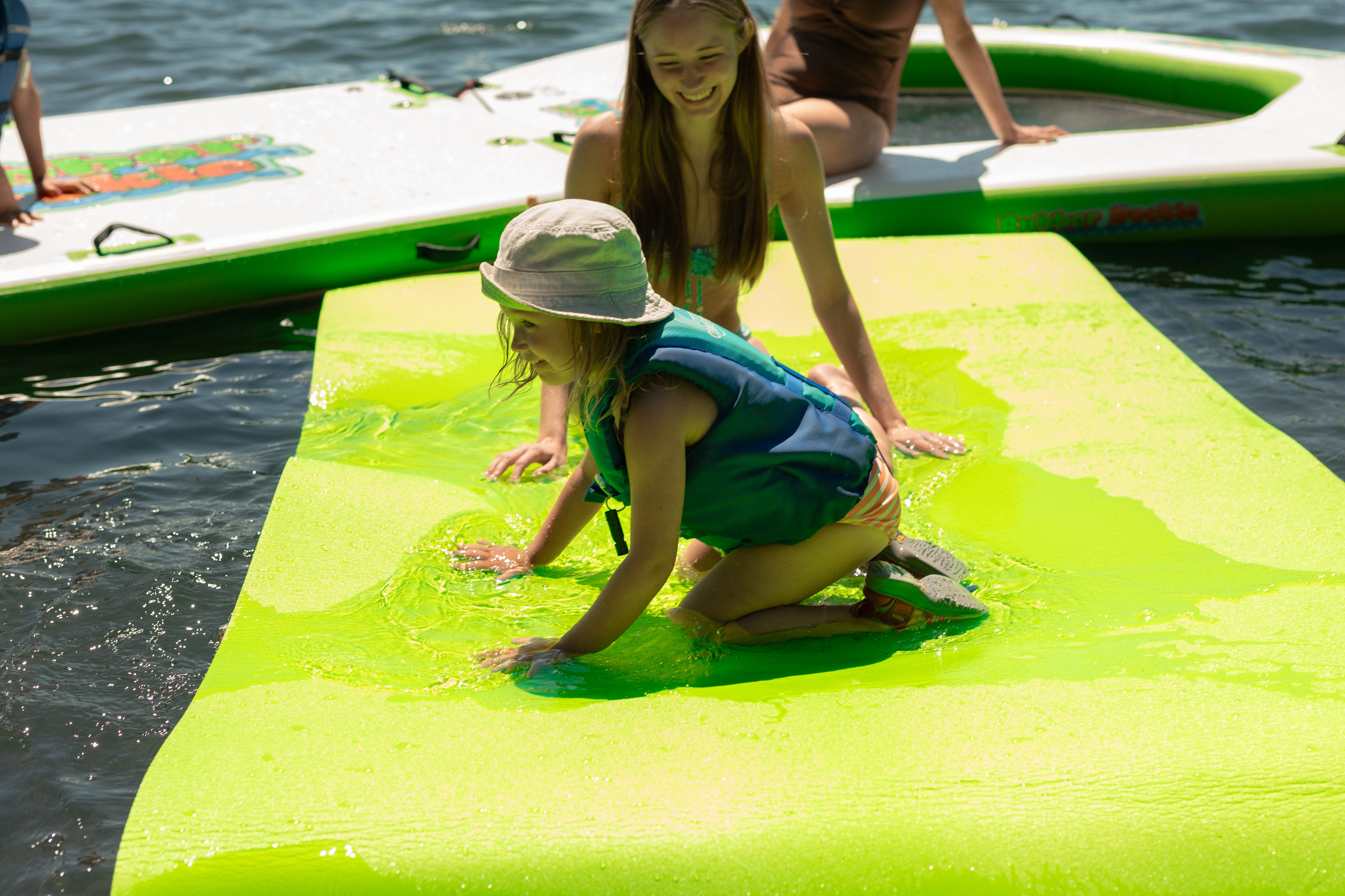 Child wearing a life jacket playing on a green Rubber Dockie 9x5 floating water mat with family on the lake