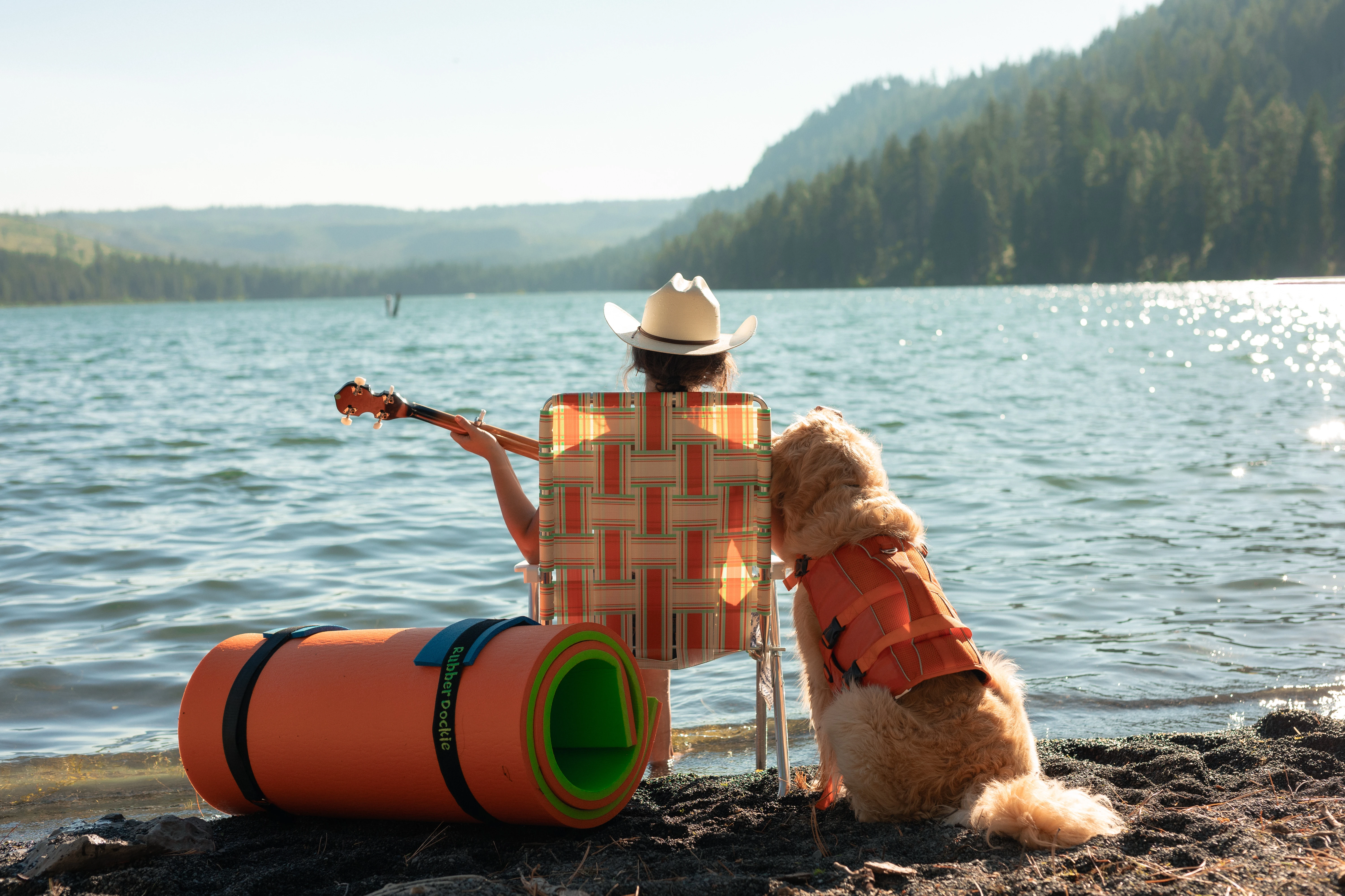 Woman playing guitar beside a dog in a life jacket and a rolled orange Rubber Dockie floating water mat on a lakeshore