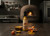 Bottle of Vicious V Curry Turmeric Hot Sauce displayed on a rustic table with turmeric root, curry powder, peppers, and a bowl of golden sauce in front of a wood-fired brick oven.