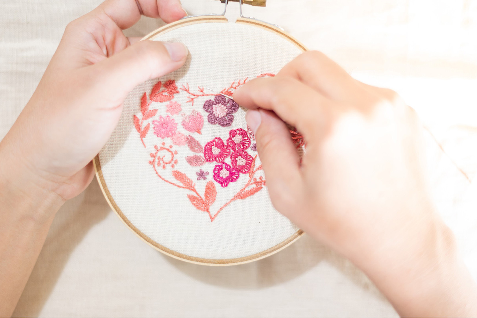 Hands embroidering a floral heart design on fabric in an embroidery hoop.