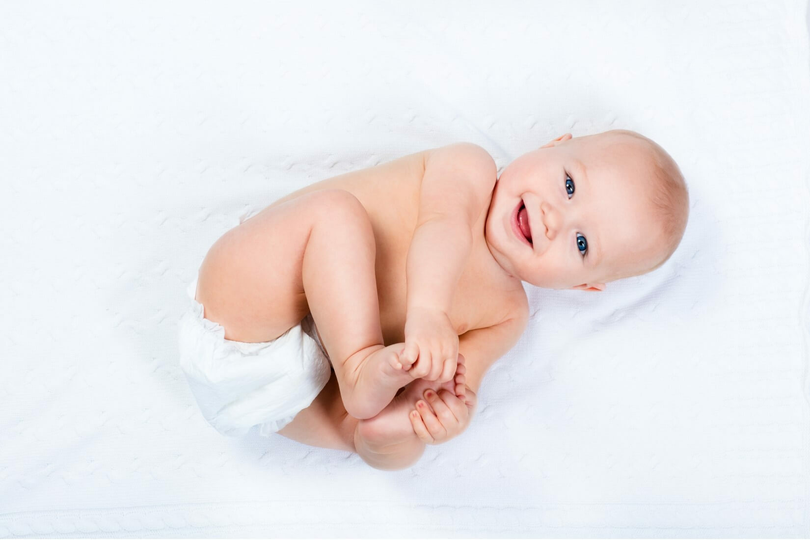 A baby lying on a white blanket, wearing a diaper, with feet and hands in focus.