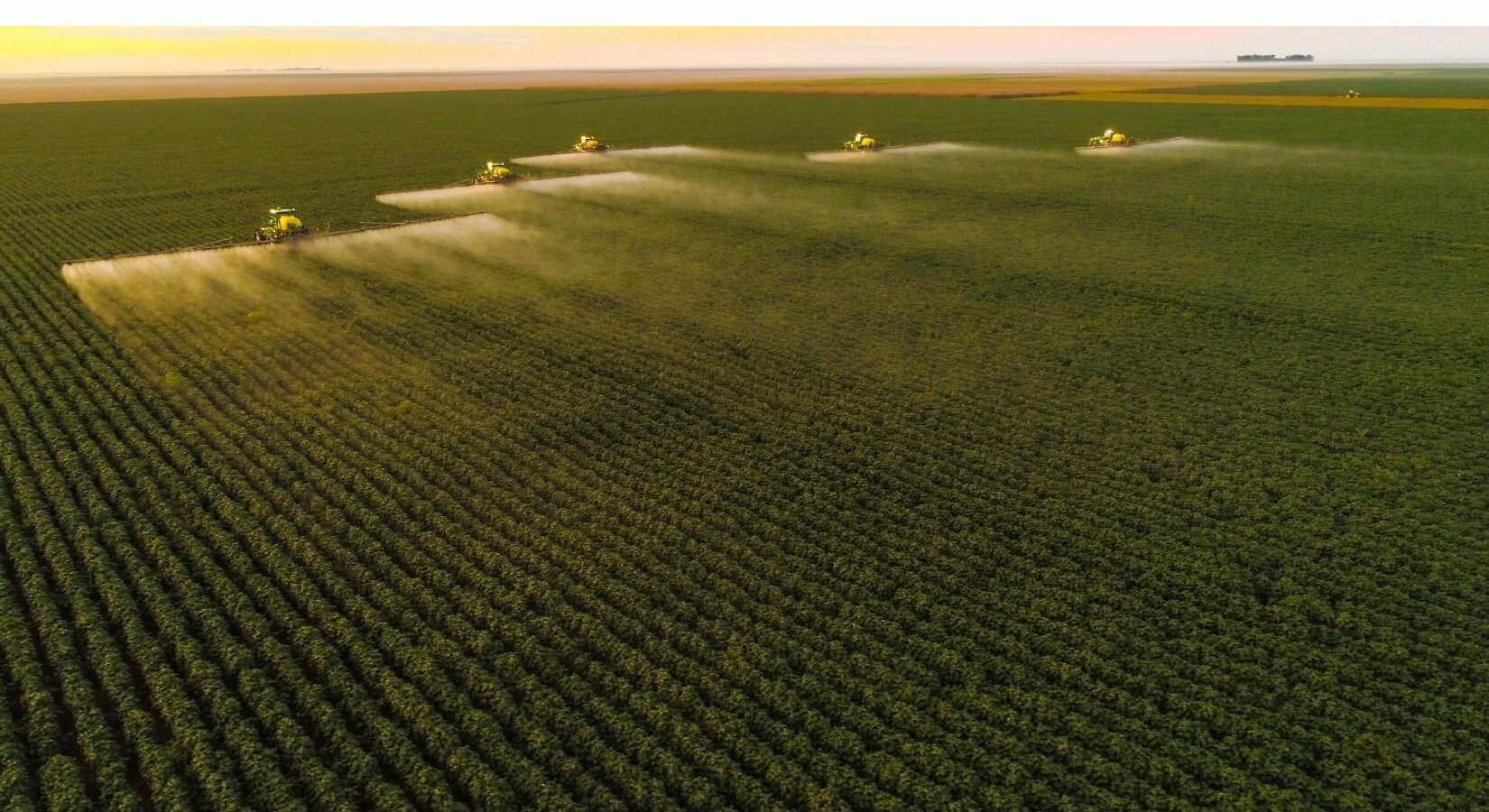 Aerial view of tractors spraying crops in a vast agricultural field at dusk.