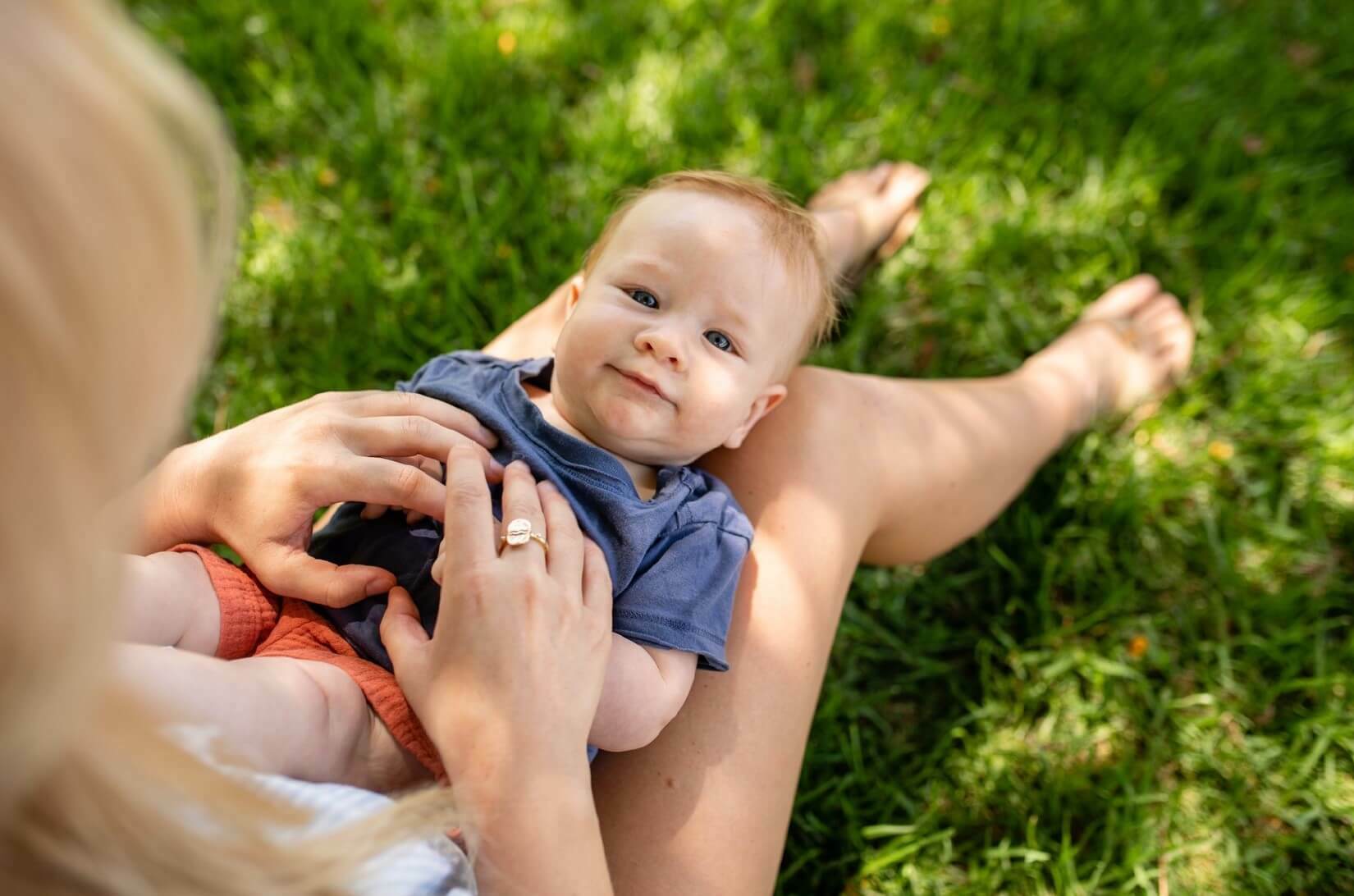 A toddler sitting on grass with an adult's hands gently touching the child's.