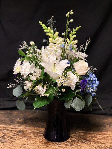  Lovely white arrangement accented with blue hydrangea, white roses, white snap dragon and additional complimentary blossoms in a blue vase