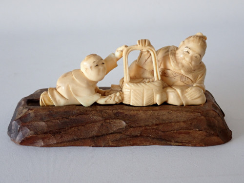 Antique Japanese Meiji period ivory okimono depicting a man and young boy beside a woven basket, signed and displayed on a carved wooden stand.