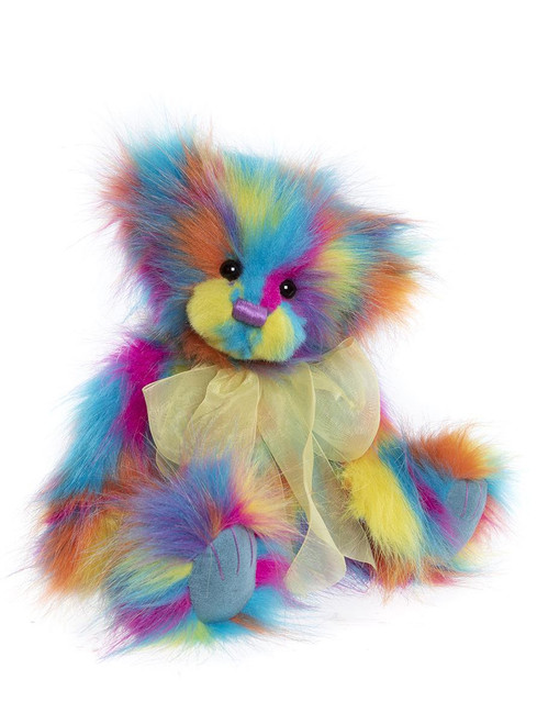 A multicoloured rainbow teddy bear sits upright against a white background, wearing a sheer yellow organza bow ribbon around its neck.