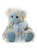 A soft plush teddy bear sits upright against a white background. It features blue and cream mixed fur, blue suede paw pads, and a pale blue organza ribbon bow at its neck.