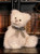 A white fluffy Charlie Bears teddy bear sits upright, wearing a grey gingham bow tie. It has silver-tipped fur, a grey velvet nose, and detailed paw pads.