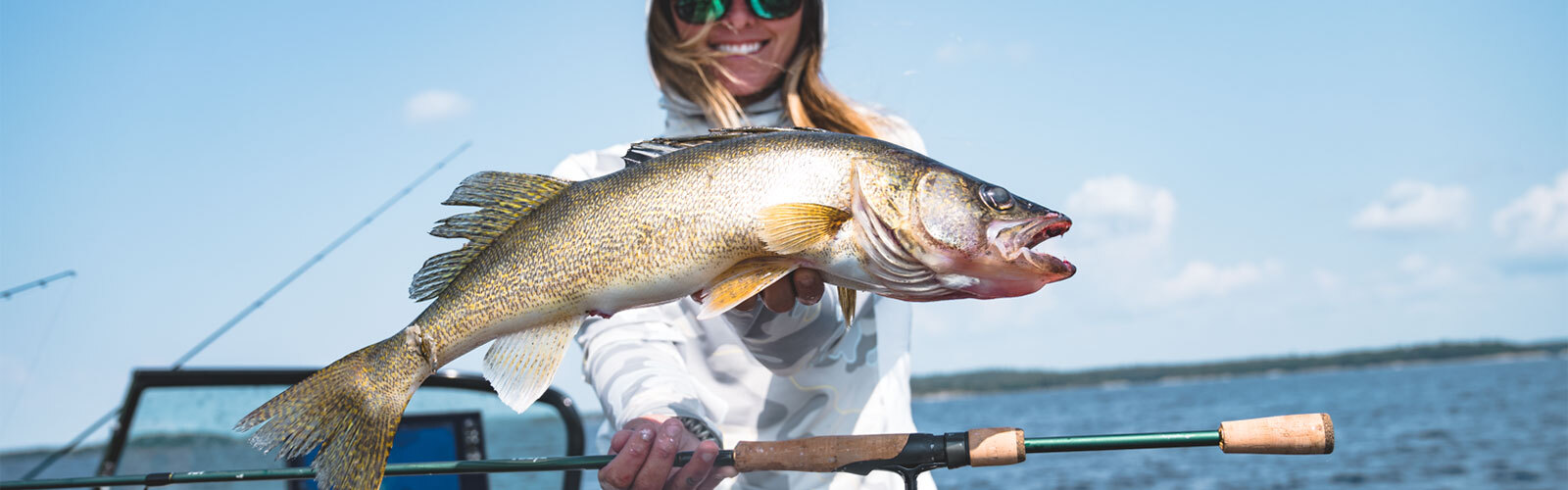 Person holding a large fish and fishing rod on a boat with a lake and clear blue sky in the background.