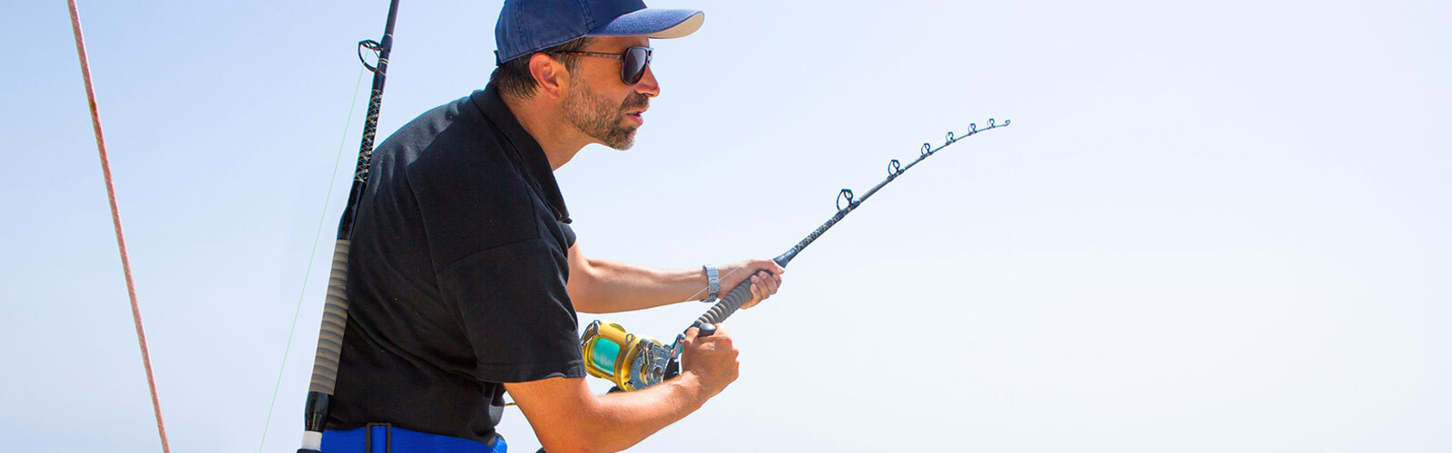 Man fishing on a boat, holding a bent fishing rod against a clear blue sky background.