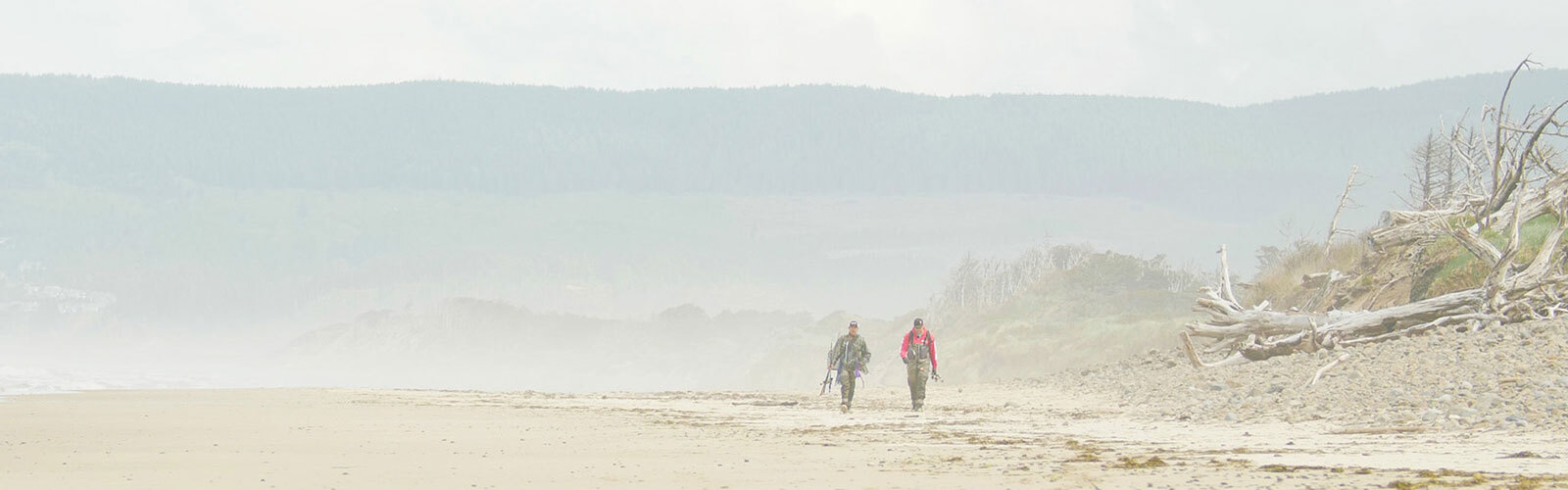 Two people walk a misty empty beach with driftwood, calm sea, and hazy forested hills in the background.
