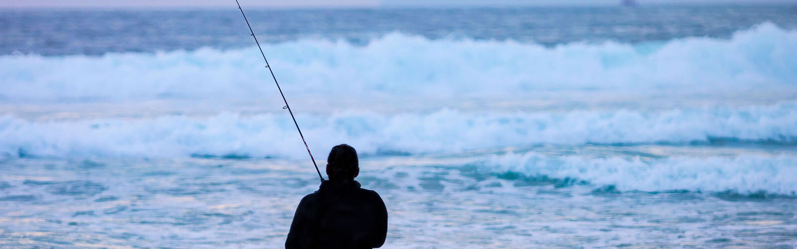 Silhouette of a person fishing at dusk, pink-purple sky above calm waves in a serene ocean scene.

