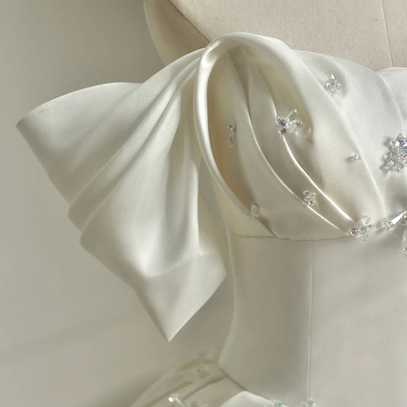 Close-up of the white satin off-the-shoulder wedding dress showing sculptural folded sleeve draping and crystal floral beading