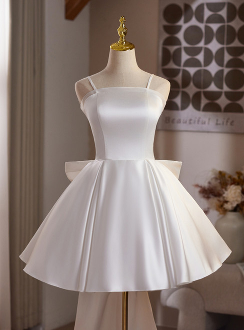 White satin A-line homecoming dress with thin spaghetti straps, a flared knee-length skirt, and a statement bow detail at the natural waist.