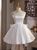 White satin A-line homecoming dress with thin spaghetti straps, a flared knee-length skirt, and a statement bow detail at the natural waist.