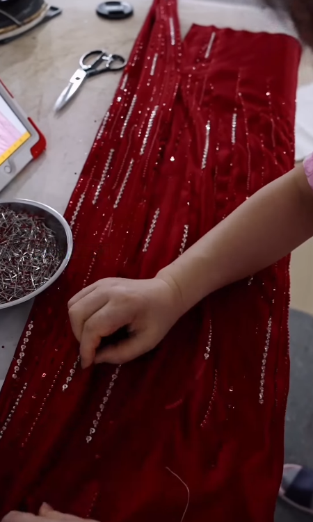 Close detail of an artisan hand sewing individual bead rows onto a red dress panel