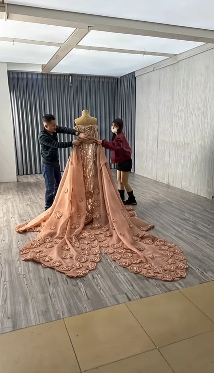 Assistants carefully arranging the voluminous detachable train of a champagne pink A-line prom gown in the atelier