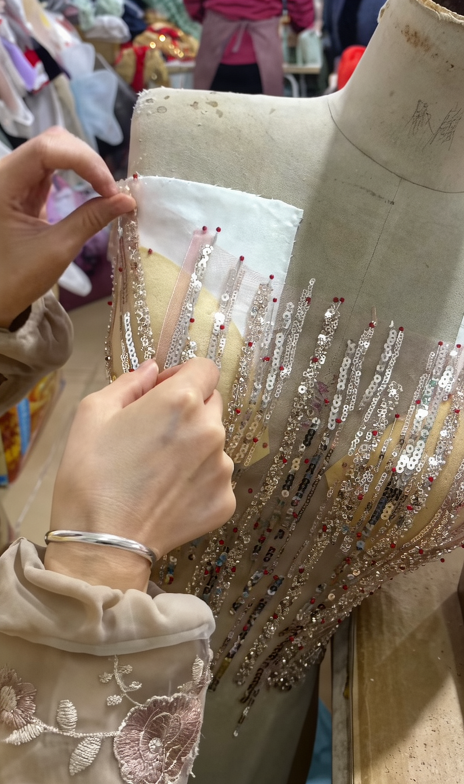 Close-up of an artisan hand-sewing champagne pink sequins and glass beads onto the bodice of the gown on a mannequin